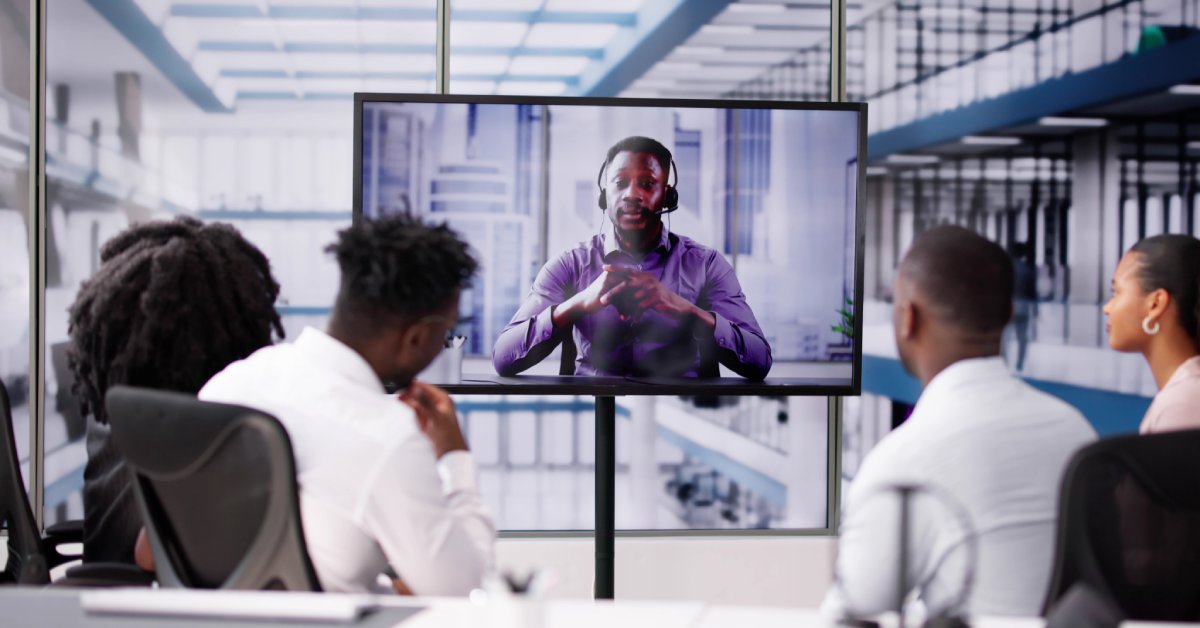 A group of people in business attire watching a screen showing a businessman wearing a headset and speaking.