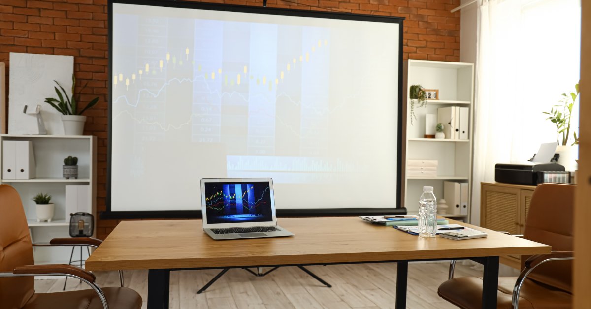 A cozy workspace with a projector screen and a table. The table has notepads, a water bottle, and a laptop on it.