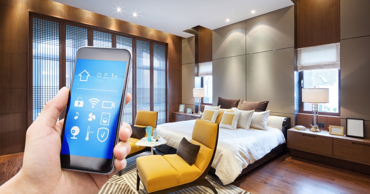 A person holding a smartphone with graphics for smart home controls. Behind the phone is a bedroom with ambient lighting.