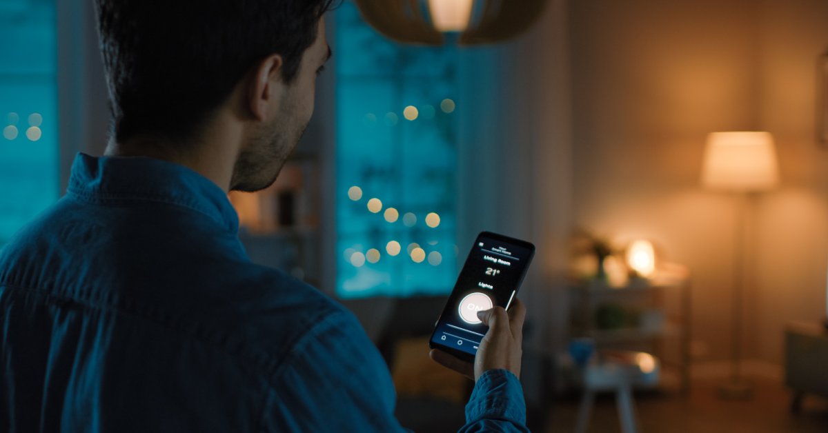 A man using a control on a smartphone while looking at a floor lamp across the room. The lighting in the room is dim.