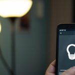 A person holding a phone displaying a graphic of a light bulb. Behind the phone is a floor lamp with two lights.