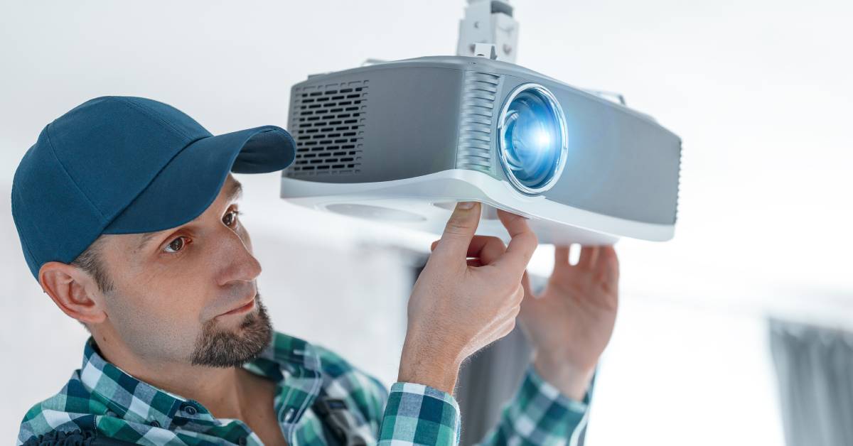 A professional installer adjusting the focus of a video projector mounted on the ceiling while looking into the distance.