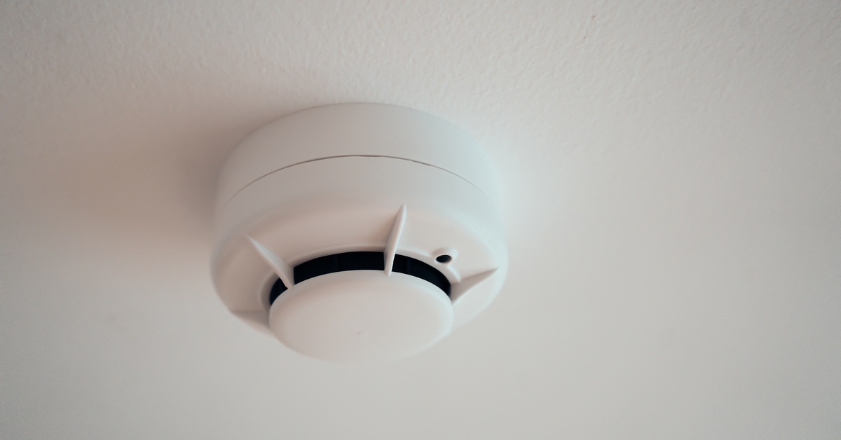 A close-up view of a white smoke detector mounted on a white ceiling. It has a circular design and a vent.