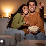 A young woman smiles, leaning on a young man as they sit on a couch. He holds a bowl of popcorn and points ahead.