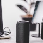 Modern desk setup with a black cylindrical speaker, smartphone on a stand, laptop, glasses, and a plant.