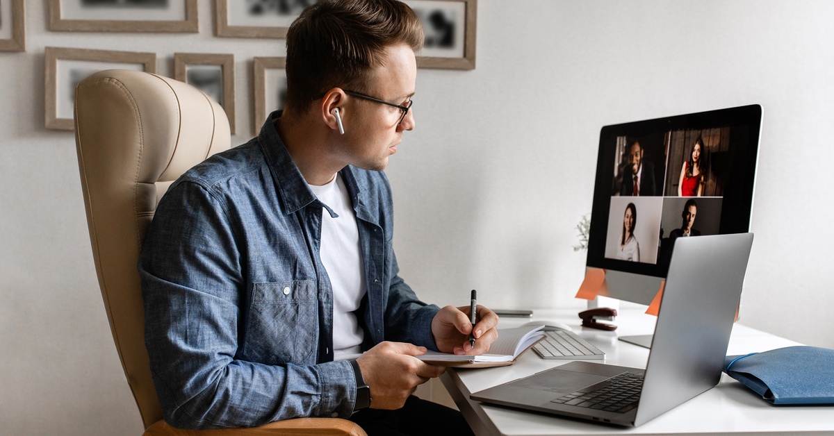 A man with glasses and earbuds sits at a desk, taking notes during a video call, with a laptop nearby.