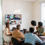 A diverse group sits at a conference table in a bright office, engaging with participants on a large screen.