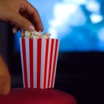 A close-up of a classic popcorn bucket as a person's hand reaches in. A television glows in the blurred background.