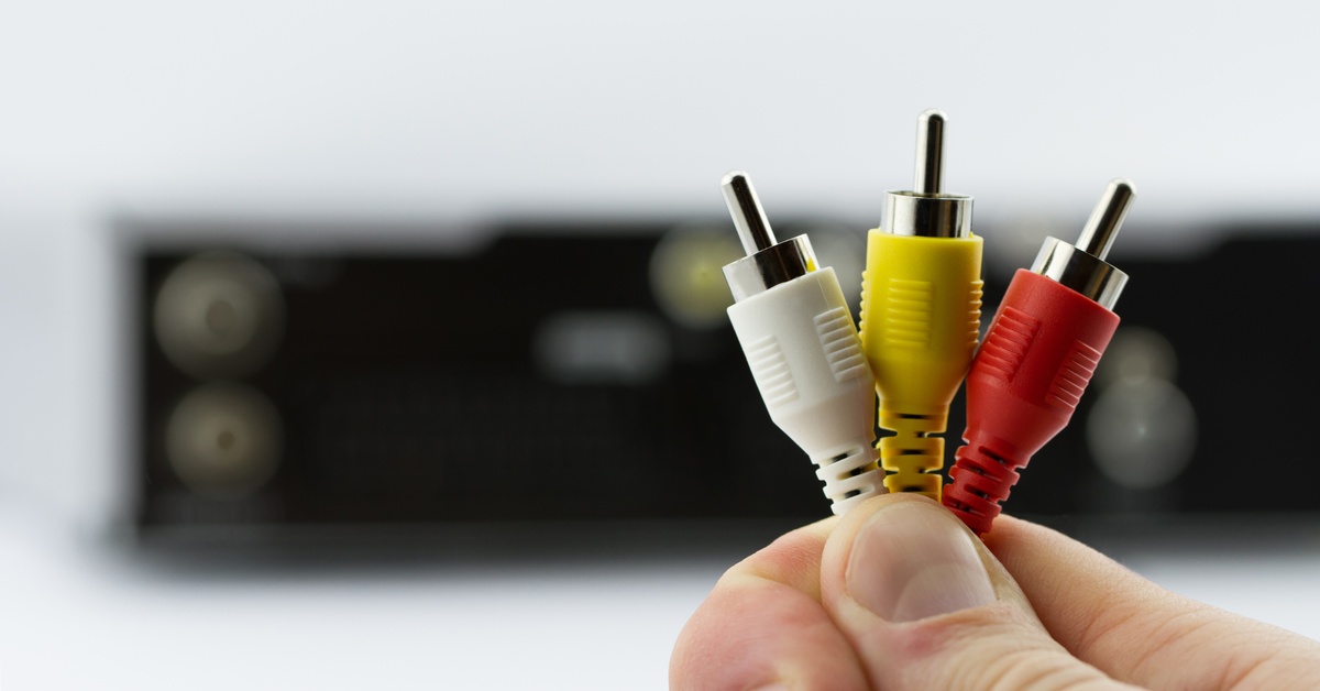 A close-up of a hand holding red, white, and yellow RCA connectors in front of blurred AV receiver equipment.