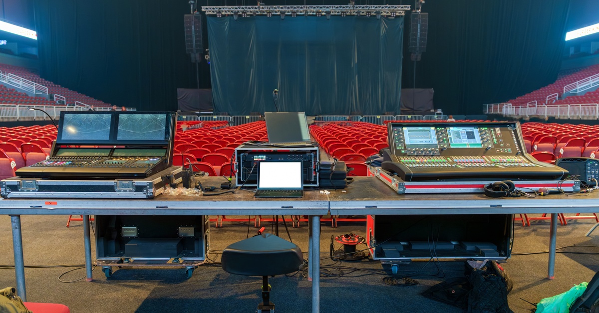 Two soundboards and a control box are arranged on a table inside a concert venue with rows of seats in the background.