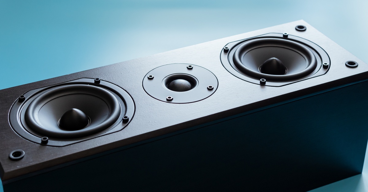 A close-up of a black loudspeaker placed on a light blue background. Soft lighting reflects off the surface.