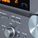 A close-up image of an AV receiver with various buttons and knobs. The digital display shows the audio channel levels.