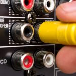 A close-up of a person connecting a yellow audio cable into input jacks on the back of electronic equipment.