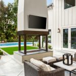 A covered patio beside a swimming pool with cushioned chairs, a wall-mounted screen, trees, and sunlight.