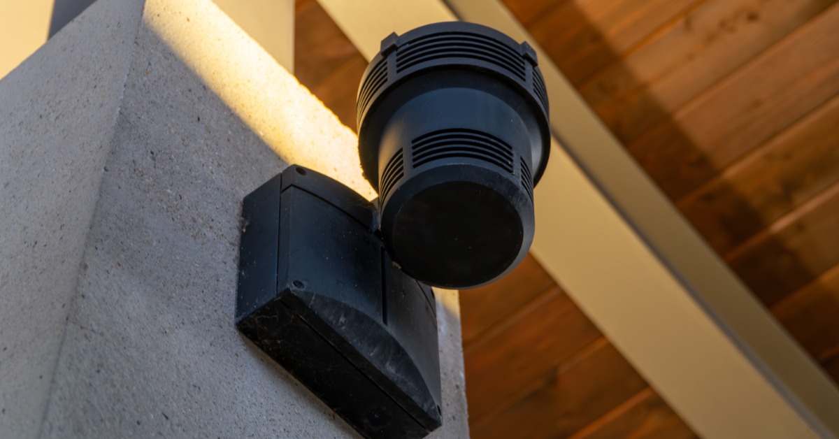 A black speaker mounted on a concrete pillar beneath a wooden ceiling with warm sunlight across the surface.