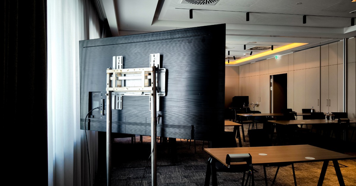 A large flat-screen monitor mounted on a bracket near windows and beige curtains in a modern conference room.