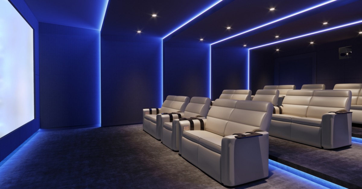 A home theater room with a large screen, beige leather armchairs, blue lighting, dark walls, and a patterned rug.