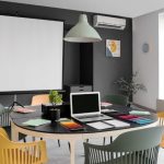 A conference room contains a round table, white chairs, a projector screen, ceiling lights, and dark walls.