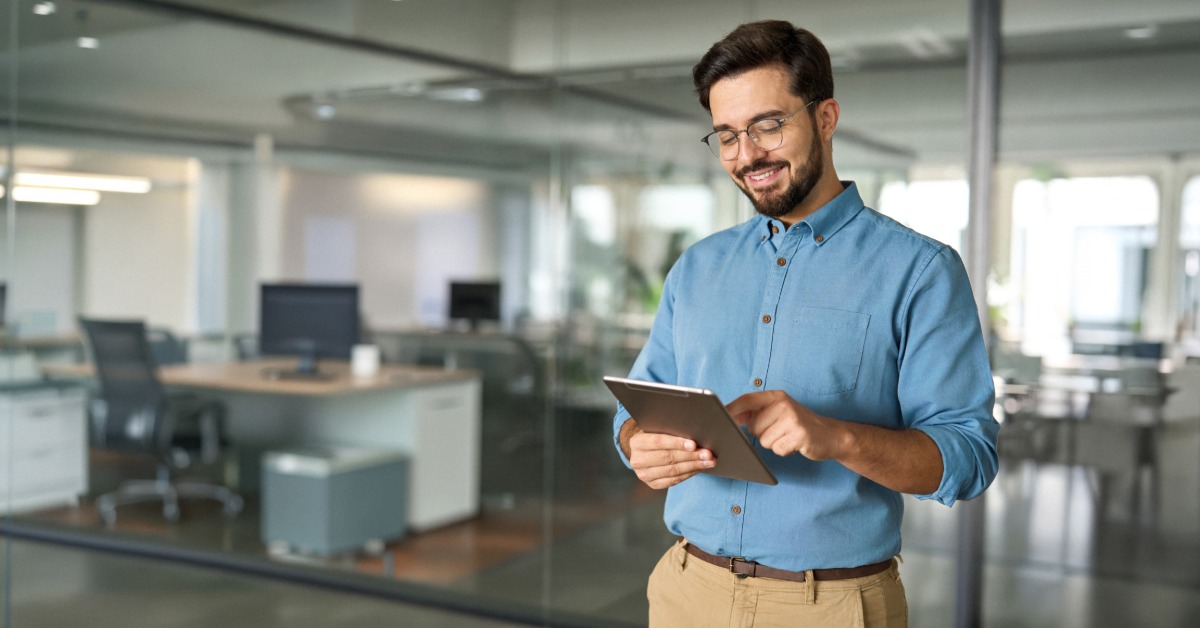 A man in professional attire stands in an office holding a tablet in front of large windows with cubicles behind him.
