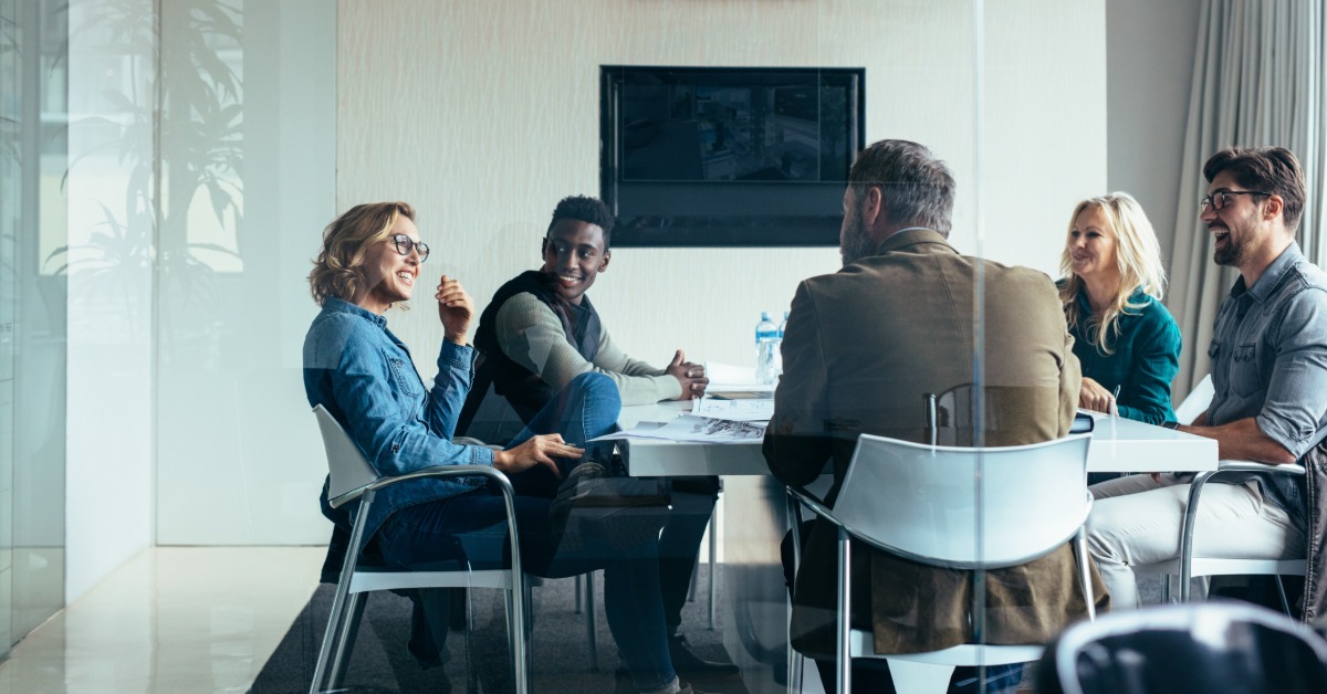 A diverse group of people sits at a table in a brightly lit conference room with a digital screen on the wall.