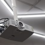 A white projector mounted to a ceiling pole among bright strip lights, gray panels, and visible cables.