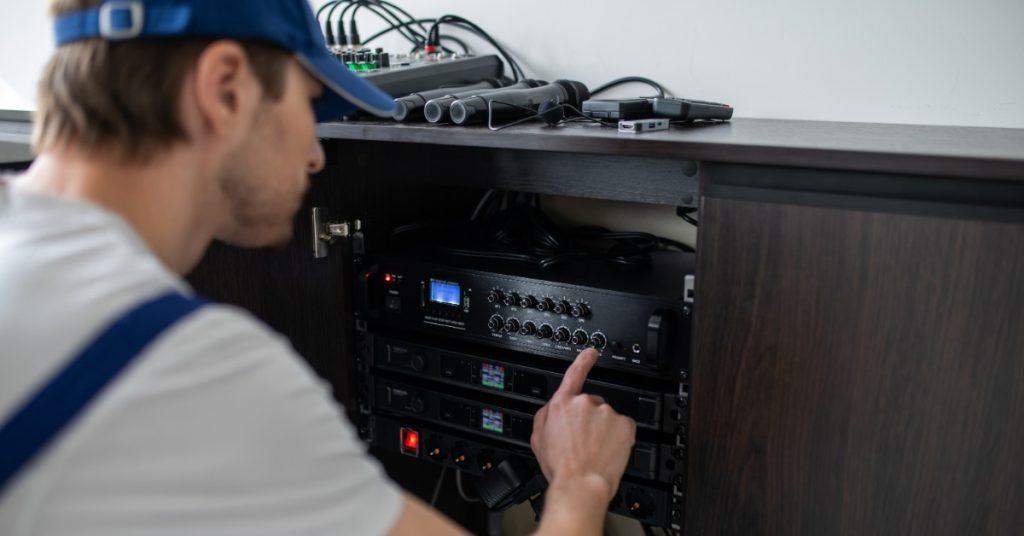 A person in a blue cap reaches toward audio equipment beneath a shelf with microphones and cables nearby.