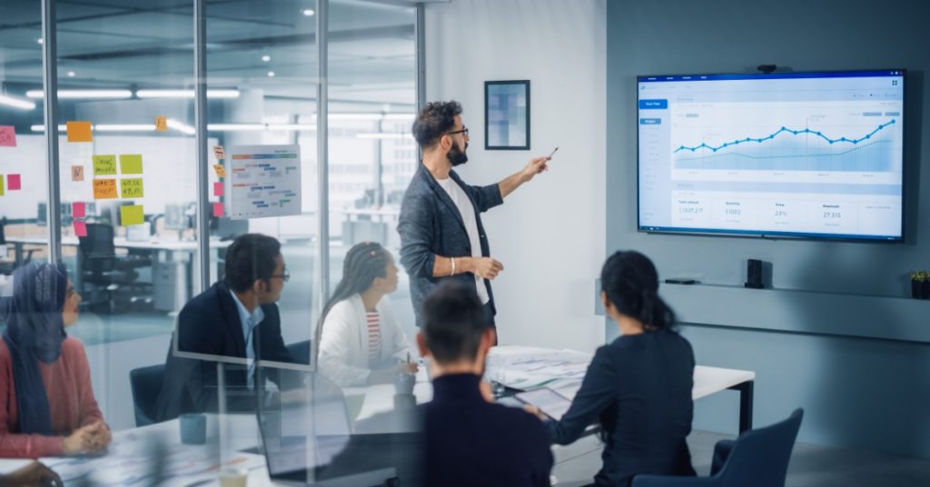A close-up of a person pointing at a wall screen with charts while coworkers sit around a table in a glass office.