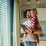 A man is holding a young child and a tablet by a large window with horizontal blinds in a bright kitchen.