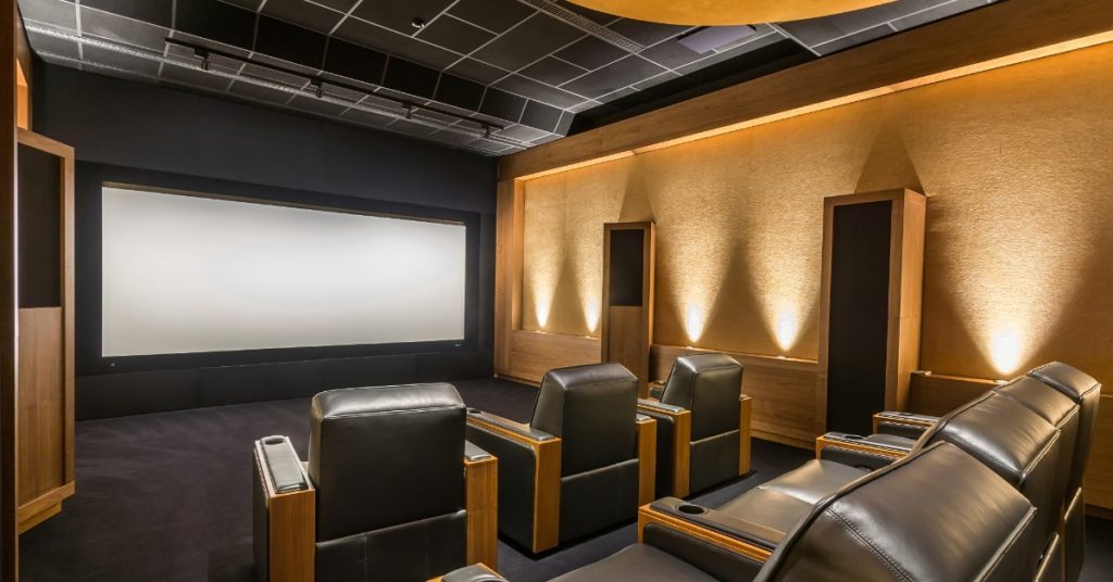 A home theater with black leather reclining chairs facing a large white screen. Warm wall lights illuminate the room.