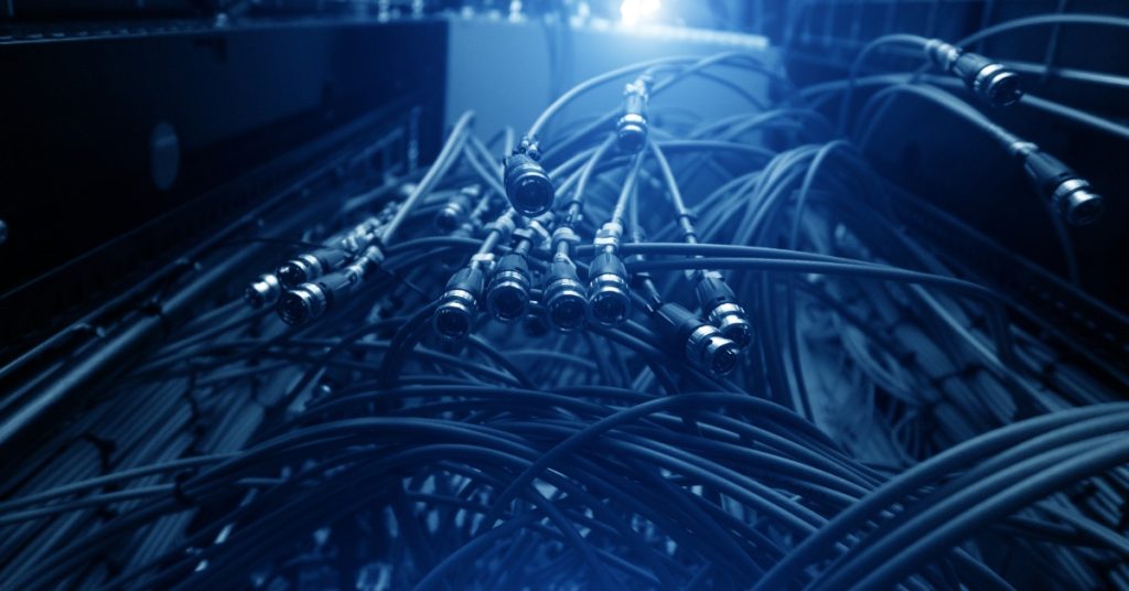 A tangled group of cables with silver metal connectors hanging inside a dark equipment rack under blue lighting.